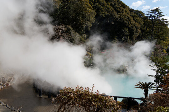 Massive billows of thick white geothermal steam rising dramatically from a turquoise hot spring pool surrounded by lush forest and paved walkway under bright sunlight.