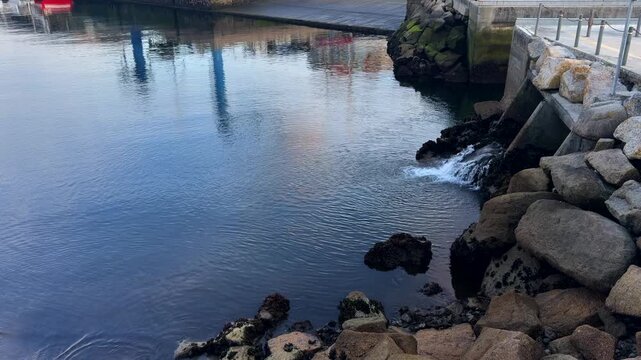 Water flowing from a drainage outfall into calm harbour water beside granite riprap and quay
