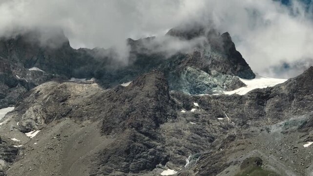 Drone flies above an extremely rugged and rocky Alpine mountain landscape with visible landslides, scree slopes and towering peaks disappearing into moody clouds under dramatic weather.