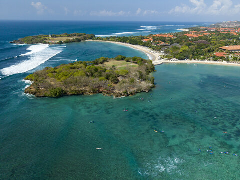 Aerial view of Nusa Dua Beach with surfers in the turquoise water, lush green vegetation on the headland, and resort buildings along the coastline in Kecamatan Kuta Selatan, Bali, Indonesia.