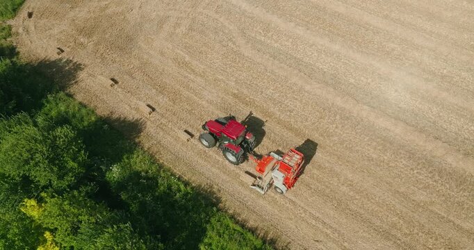 Top-down drone view of a straw baler opening its rear gate to release a square straw bale on a harvested wheat field, framed by tree-lined edges and striking geometric field patterns.