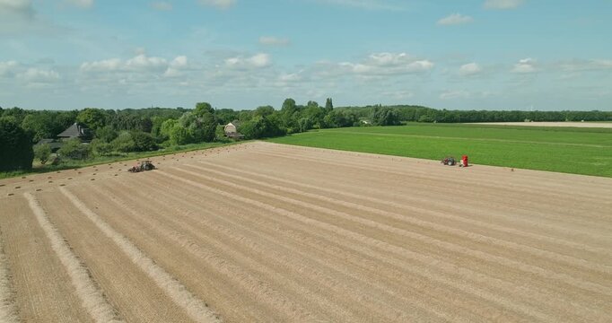 Drone flies toward a red tractor towing a straw baler across a harvested wheat field, producing straw bales beneath a clear blue summer sky in the Dutch countryside.
