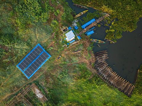 Aerial view of Lake Rawa Pening featuring a floating solar panel array, wooden boats moored along the grassy shore, and lush vegetation in Waduk Rawapening, Central Java, Indonesia.