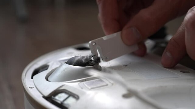 Male hands using maintenance tool with blade to cut and remove clump of tangled hair and dust from small front wheel of automatic robot vacuum cleaner. Concept of maintenance modern home appliance.