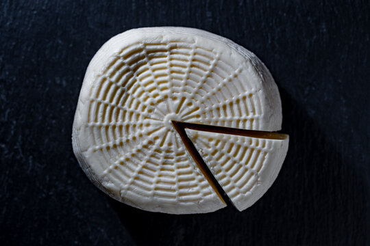 Fresh homemade goat cheese wheel on black background, closeup. White artisan cheese wheel with a cut slice on dark slate background top view