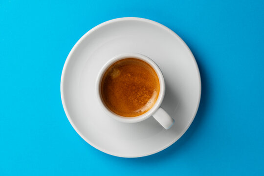Espresso coffee cup on bright blue background, closeup. Top view of fresh espresso coffee in white ceramic cup with saucer on blue background
