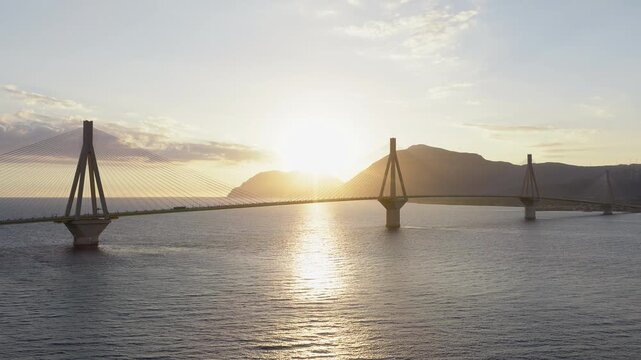 Drone pan of Rio-Antirrio Bridge silhouetted at sunset. Sun disappears behind pylons then re-emerges. Calm sea, golden reflections