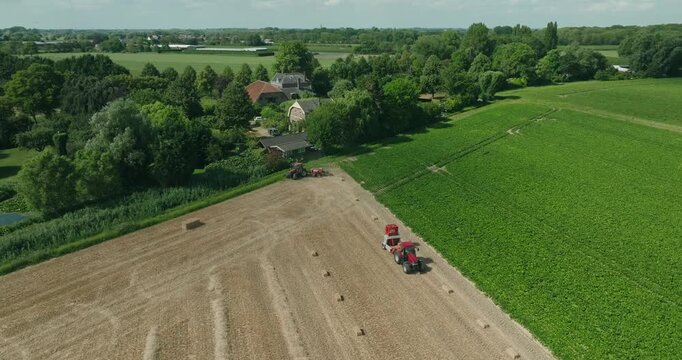 Drone orbits a tractor baling straw across harvested wheat fields, surrounded by the picturesque Dutch countryside beneath a clear blue summer sky with scenic farmland.