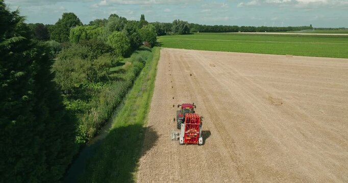 Drone follows a tractor from behind as it collects square straw bales across a harvested wheat field beneath a clear blue summer sky in the Dutch countryside.