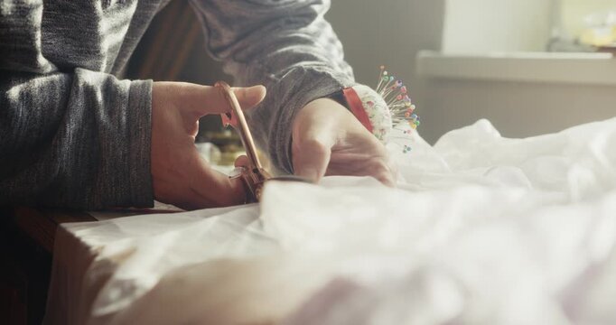 Close-up of a fashion designer cutting white fabric beside a bright window, illustrating dressmaking, textile craftsmanship and creative small business work.