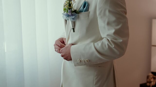 Groom in a cream suit with boutonniere and blue pocket square fastening his cuff by a window