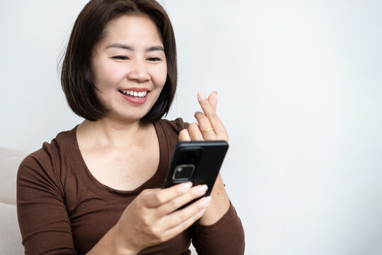 Happy Asian woman showing mini heart sign while video calling on smartphone, representing romance scam, online dating, digital love, virtual relationship, and mobile chat