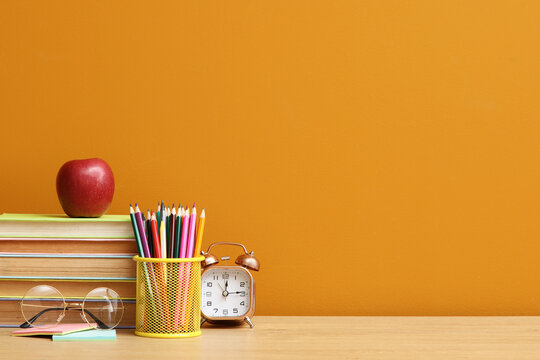 School supplies with a red apple, books, pencils, and an alarm clock on a wooden desk against an orange wall