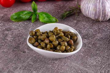 Fototapeta premium Capers in white bowl with basil garlic and cherry tomatoes on textured surface