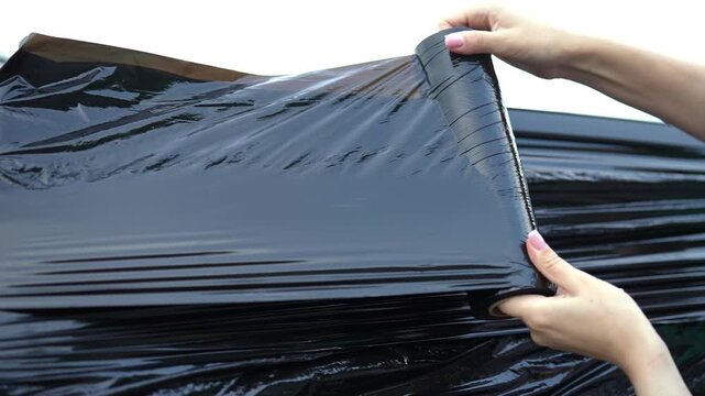 Close-up of female hands unrolling black stretch film. Woman covering vehicle with protective plastic wrap. Pallet wrap used for packaging, shipping, as prank. Outdoor setting, closeup, slow motion.