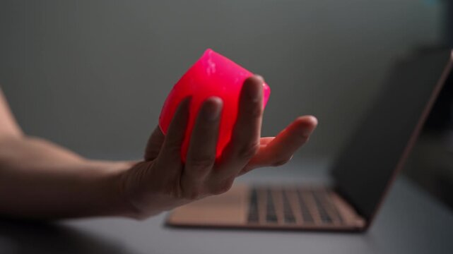 Close-up person hand squeezing vibrant pink stress relieving slime ball at modern office desk, managing anxiety and promoting relaxation through sensory fidgeting working. Shooting in slow motion.
