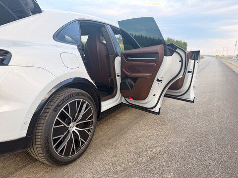 White sedan car parked on the roadside with rear doors open, showing a luxury interior and alloy wheel on an empty highway in daylight.