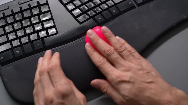 Close-up top view of person cleaning dusty ergonomic computer keyboard and wrist rest with special pink cleaning slime. Man removing dirt and dust from office equipment for hygiene, slow motion.