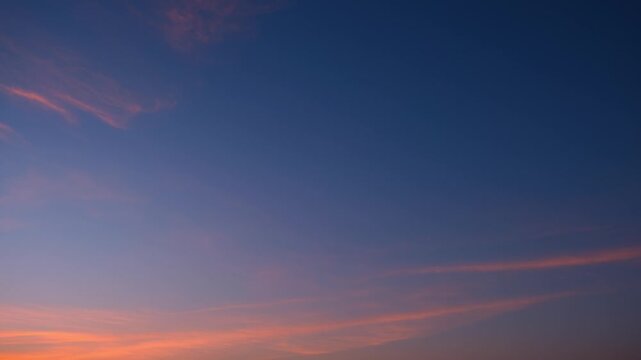 A sunrise sky timelapse video featuring wispy cirrus clouds illuminated by soft coral, pink, and orange sunlight, creating a dramatic color gradient beneath the deep blue early morning sky. 4k.
