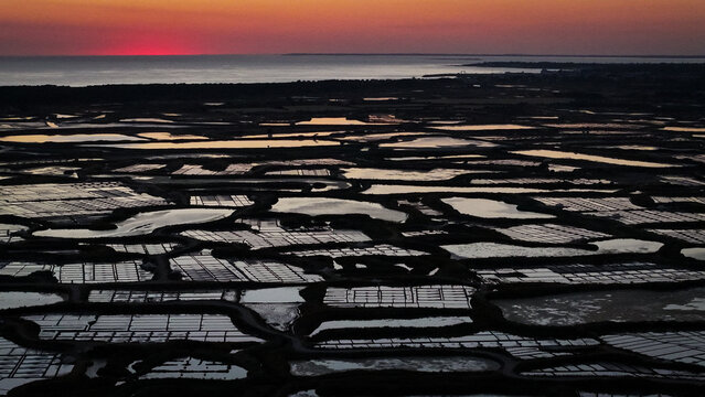 Aerial view of the Marais salants de Guerande at sunset, featuring reflective salt pans and dikes under a vibrant red sky in Guerande, Pays de la Loire, France.