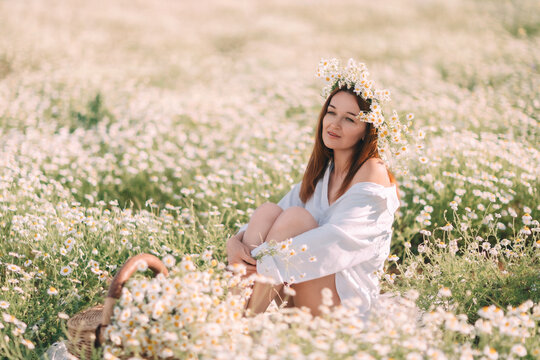 Peaceful woman sitting in meadow of Matricaria chamomilla wearing white shirt and floral crown during sunny spring day
