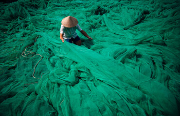 Naklejka premium Vietnamese fisherman with conical hat mending green fishing nets outdoors, traditional craft