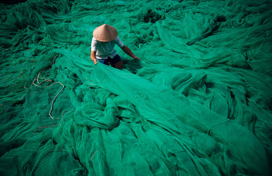 Vietnamese fisherman with conical hat mending green fishing nets outdoors, traditional craft