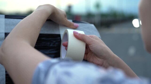 Close-up of young female hands applying white masking tape to black plastic garbage bag, creating makeshift temporary repair for broken or shattered car window after unfortunate incident, slow motion.