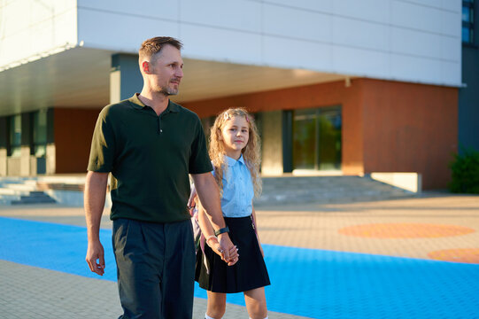 A father holding hands with his young daughter walking to school on a sunny day