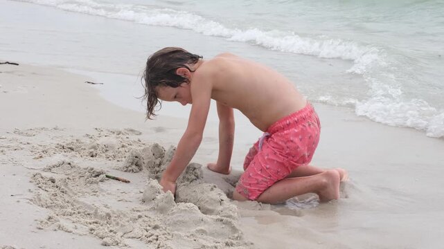 A young boy kneels on the shoreline while digging and shaping wet sand beside gentle ocean waves. The candid beach scene highlights childhood creativity, outdoor recreation, and imaginative play.