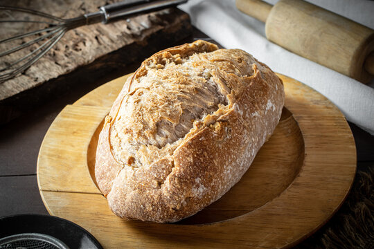 Freshly baked artisan sourdough bread loaf resting on a wooden board in a rustic kitchen setup with flour, a rolling pin, and traditional baking equipment. 