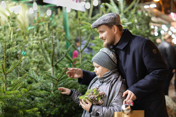 Obraz premium During Christmas market, dad and son choose Christmas tree and try on balls for New Year tree. They buy Christmas tree to decorate house and apartment, preparing for New Year and Christmas.