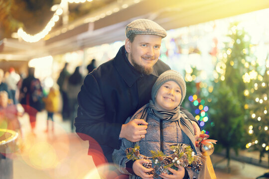 Boy with father stand in middle outdoor New Years shopping malls, dreamily imagines Christmas holiday. They stared at beautiful goods in window of fair, enthusiastically watching something fascinating