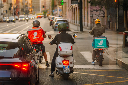 Delivery riders on scooters waiting at a traffic light in Milan, with visible food delivery bags and city streets in the background