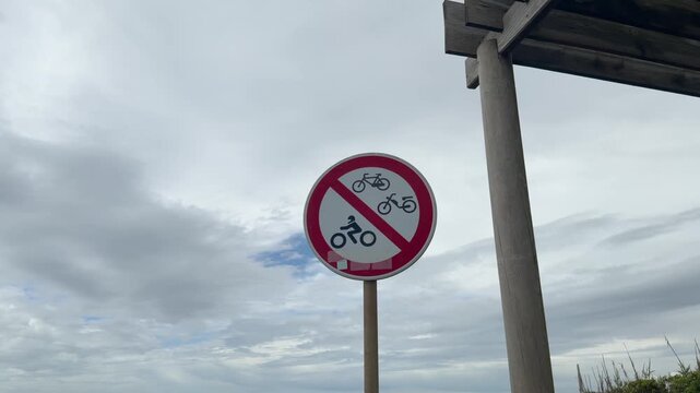 Prohibition sign banning bicycles mopeds and motorcycles beside a wooden shelter under cloudy sky