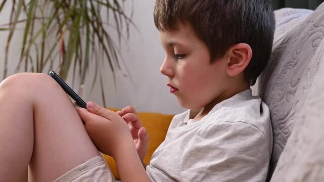 A 6-7 year old child sits on a windowsill at home with a mobile phone and plays. Mental health issues, unlimited gadgets, FOMO, harm to the child's psyche.