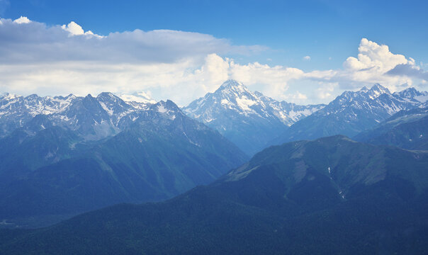Landscape of high rocky mountains focusing on a majestic central snowy peak above dark green ridges under a blue sky with fluffy white clouds.