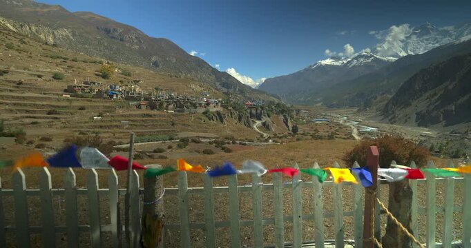 Colorful prayer flags on a fence, overlooking terraced hills, Annapurna CIrcuit, Manang Nepal, a river valley, and snow-capped Himalayan peaks