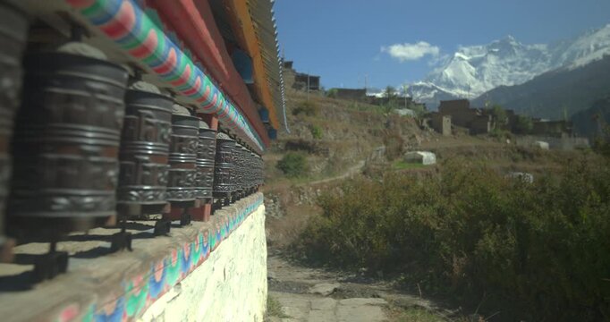 Annapurna Circuit, Manang, Nepal, gliding shot along a row of metal Buddhist prayer wheels mounted on a white stone wall, framing distant snow-covered Himalayan mountain peaks