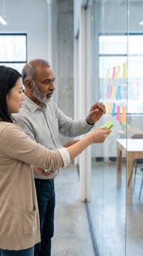 Asian professional and mature colleague planning with sticky notes on glass wall