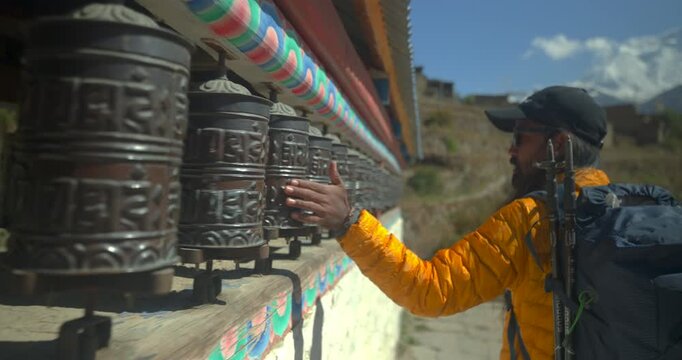 Annapurna Circuit, Manang, Nepal, tracking shot of male trekker wearing a yellow jacket and backpack as he touches and spins a row of traditional metal Buddhist prayer wheels on an outdoor stone wall.