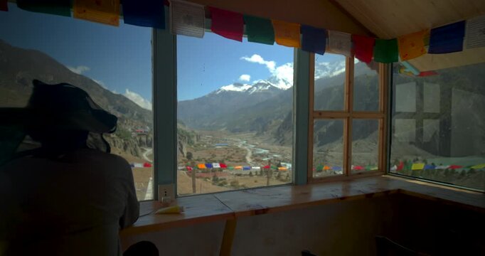 Annapurna Circuit, Manang, Nepal, gliding toward a large glass window inside a tea house, framed by prayer flags and revealing an expansive view of mountain peaks and valley floor.