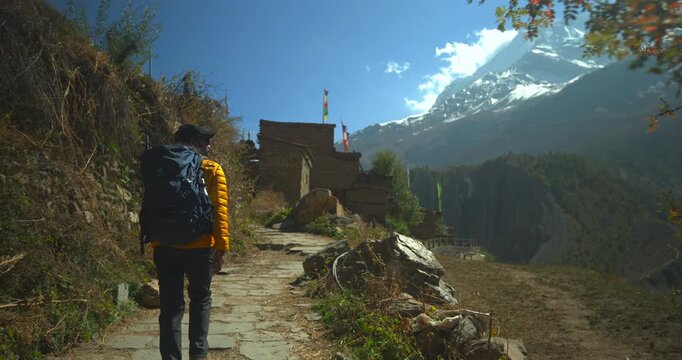 Annapurna Circuit, Manang, Nepal, tracking shot following a male trekker with a backpack walking along a trail toward stone buildings and mountain peaks.