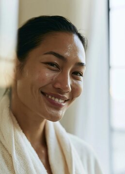 Close-up portrait of a young woman with a big smile on her face. she is wearing a white bathrobe and her hair is pulled back in a bun.
