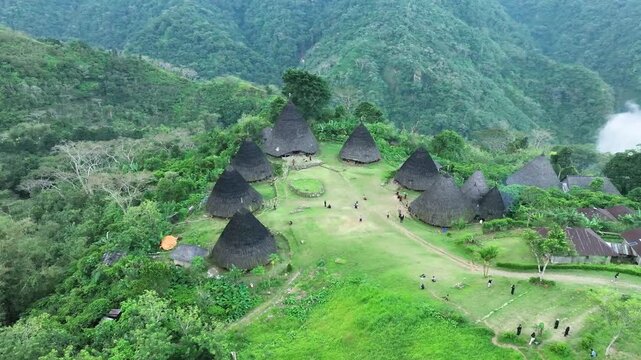 Aerial view of Waerebo Village featuring traditional conical houses set amidst lush green mountains and grassy clearings in Satar Mese Barat, East Nusa Tenggara, Indonesia.
