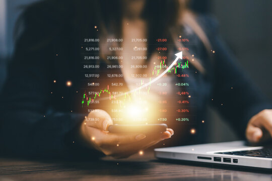 Businesswoman holding smartphone with rising stock market chart beside laptop, representing investment growth, trading, financial analytics, market performance, and digital finance.