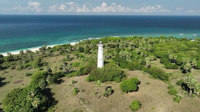 Aerial view of the Nuca Molas lighthouse surrounded by lush palm trees and coastal vegetation overlooking the turquoise sea in Satar Mese Barat, East Nusa Tenggara, Indonesia.