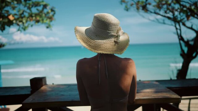 Lady relaxing in beach bar with amazing ocean view from terrace, back view. Summertime on coastline, rest in paradise, slender woman in hat admiring picturesque seascape, calm turquoise sea water