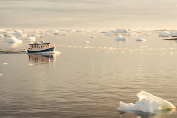 Small boat navigating through floating ice near Ilulissat, Greenland, in warm golden evening light. Peaceful Arctic seascape with icebergs and drifting ice on the calm waters of Disko Bay. © Dagmar