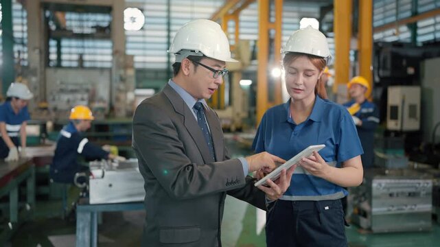 Two professionals review data in a factory setting. One wears a suit, the other a blue work uniform. Both wear helmets and focus on a tablet together
